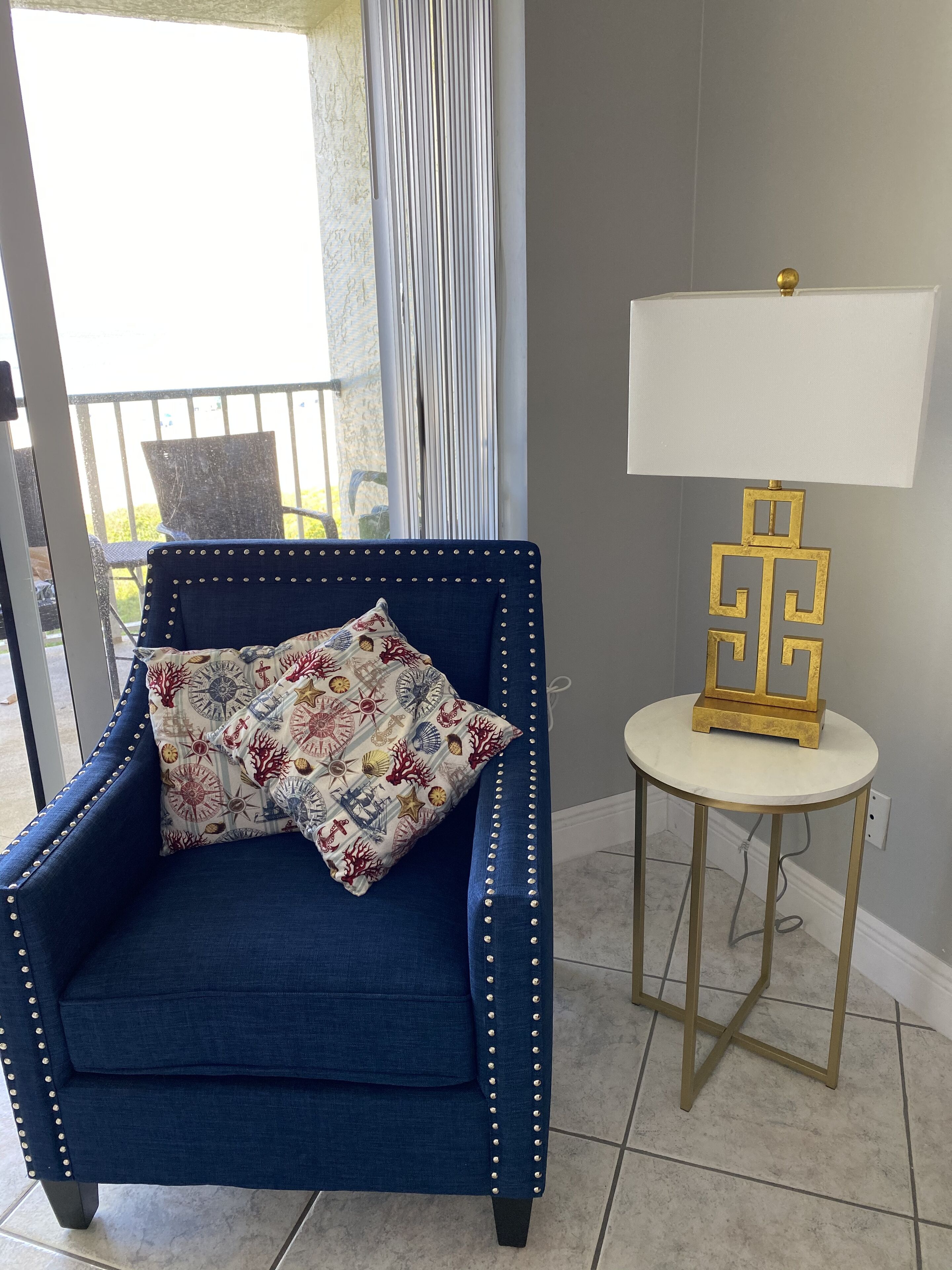 Ein blauer Sessel mit einem Zierkissen, eine goldene Lampe und ein Blick ins Freie durch ein Fenster.