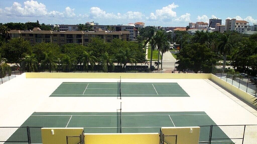 An outdoor tennis court with a net, surrounded by a yellow wall and palm trees.
