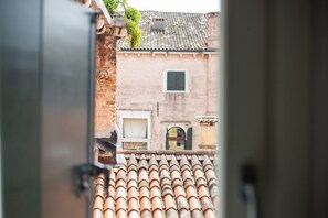 Une vue à travers une porte ouverte d’un bâtiment doté d’un toit en tuiles, d’une fenêtre avec un volet et d’un petit balcon orné d’une plante.