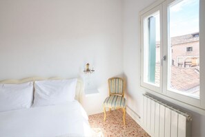 Ein Schlafzimmer mit einem Bett, einem Sessel, einem Heizkörper und einem Fenster mit Blick auf Gebäude.
