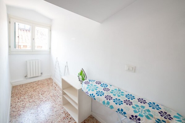 A room with a white radiator, a window with blinds, a metal shelf, and a patterned ironing board. - Ca' Rosalba, Biennale next to San Marco (Venezia)