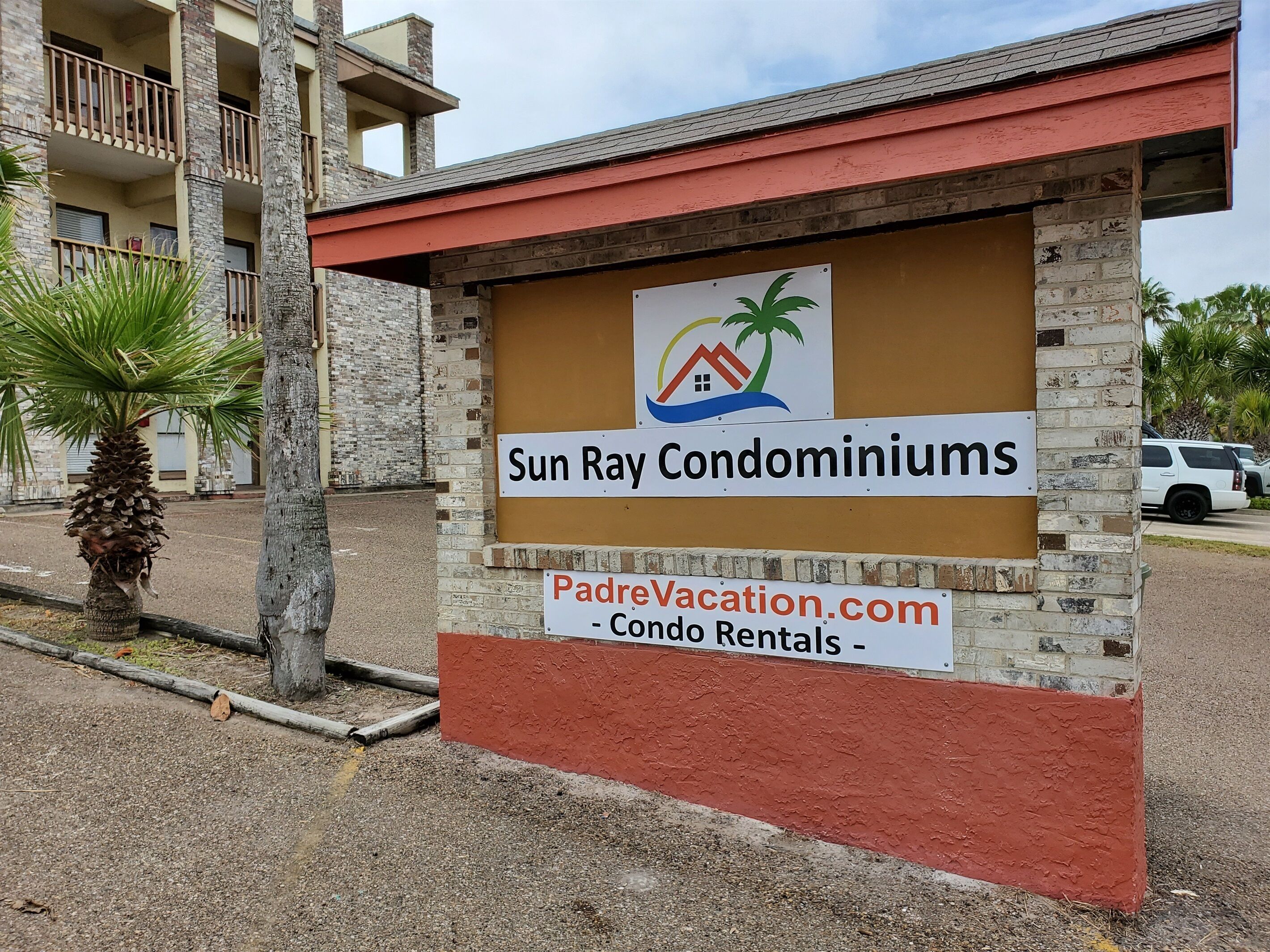 Skilt for Sun Ray Condominiums med logo, der viser et hus, en palme og en sol.