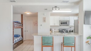 A compact kitchen with white cabinets, a microwave, and a refrigerator. There is a small bar area with two stools. - Sun Ray #3B - Pet Friendly, 3min Walk to Beach (South Padre Island)