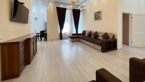 Una sala moderna con un sofá modular, un televisor de pantalla plana montado en la pared, una zona de comedor con mesa y sillas, y un candelabro.