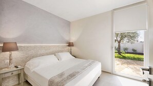 Una moderna camera d'albergo con un letto grande, due lampade da comodino, un comodino e vista su un giardino tramite una porta scorrevole.
