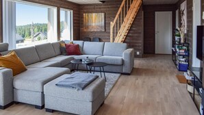 A modern living room with a grey sectional sofa, a small ottoman, and a coffee table. There is a wooden staircase leading to another level, and a large window offering a view of trees and a house. - 4 bedroom cozy home in Sjusjøen (Sjusjøen)