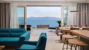 A modern living room with a sofa and chairs, a wooden dining table set for four, and a large sliding glass door leading to a balcony with a view of the sea and mountains. - LUXURY VILLA BLUE MOON (PODACA)