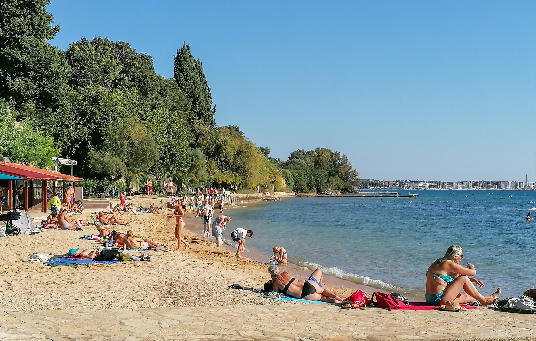 Une plage où des gens se prélassent au soleil et se baignent, une jetée en arrière-plan et un ciel dégagé.