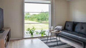 A modern living room with a large sliding glass door leading to a garden, a black leather sofa, a small round table with a remote control, and a TV on a wooden stand. - 2 bedroom amazing home in Kirke Såby (Kirke Såby)
