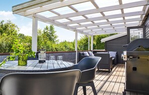 A covered outdoor patio with a table and chairs, a grill, and a view of a forested area.