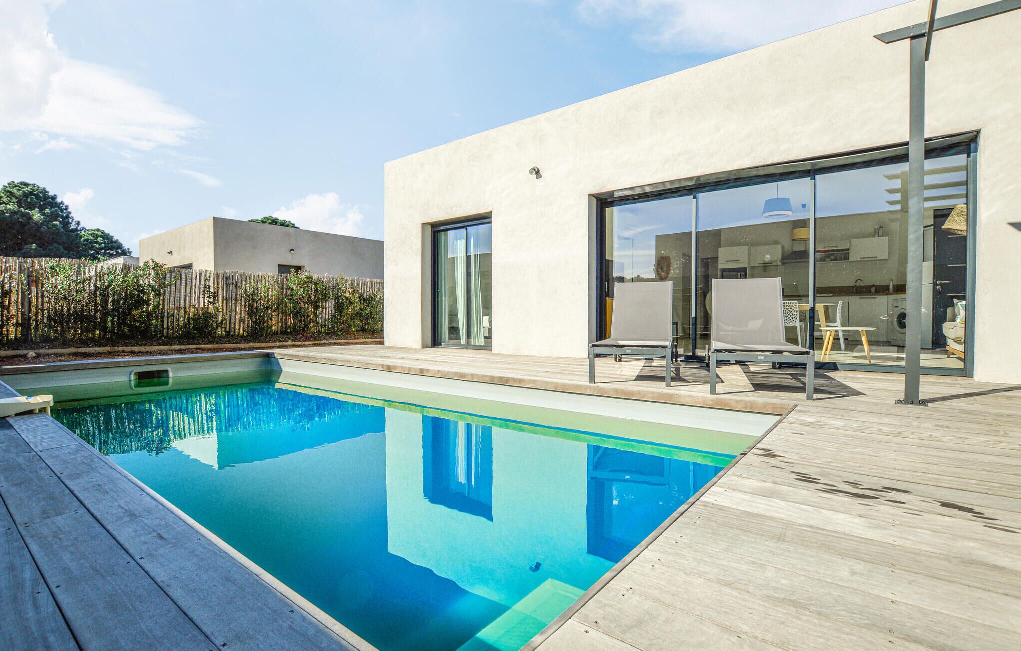 A modern outdoor pool area with a wooden deck, a clear blue pool, and a contemporary building with large glass doors and a kitchen visible inside.