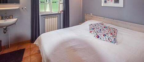 A bedroom with a bed, a bedside table, a window with curtains, a radiator, a wall-mounted light, and a framed picture.