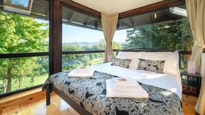 Ein Schlafzimmer mit einem großen Bett, zwei Kissen und Blick auf eine grüne Landschaft durch große Fenster.