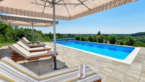 Ein Schwimmbecken mit Liegestühlen und Sonnenschirm, umgeben von einer gefliesten Terrasse und viel Grün.