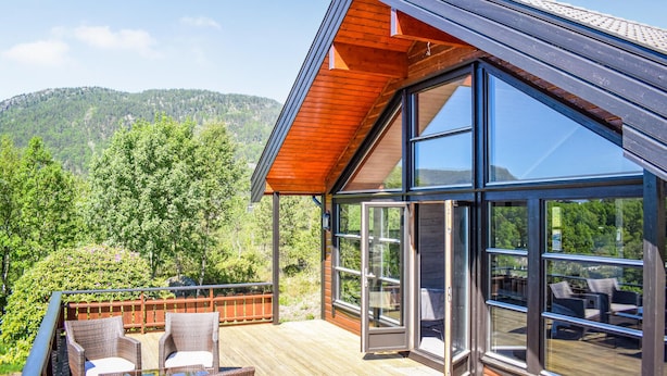 Ett modernt hus med stora glasfönster, en träveranda och utsikt över ett skogsbeklätt berg.