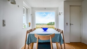 Un coin repas avec une table dressée pour quatre personnes, une vue sur un champ par une grande fenêtre et une porte sur la droite.