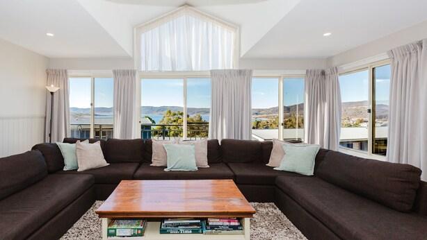 A living room with a sectional sofa, a coffee table, and large windows offering a view of a landscape. - Torino 7 - 7/14 Nettin Circuit (Jindabyne)
