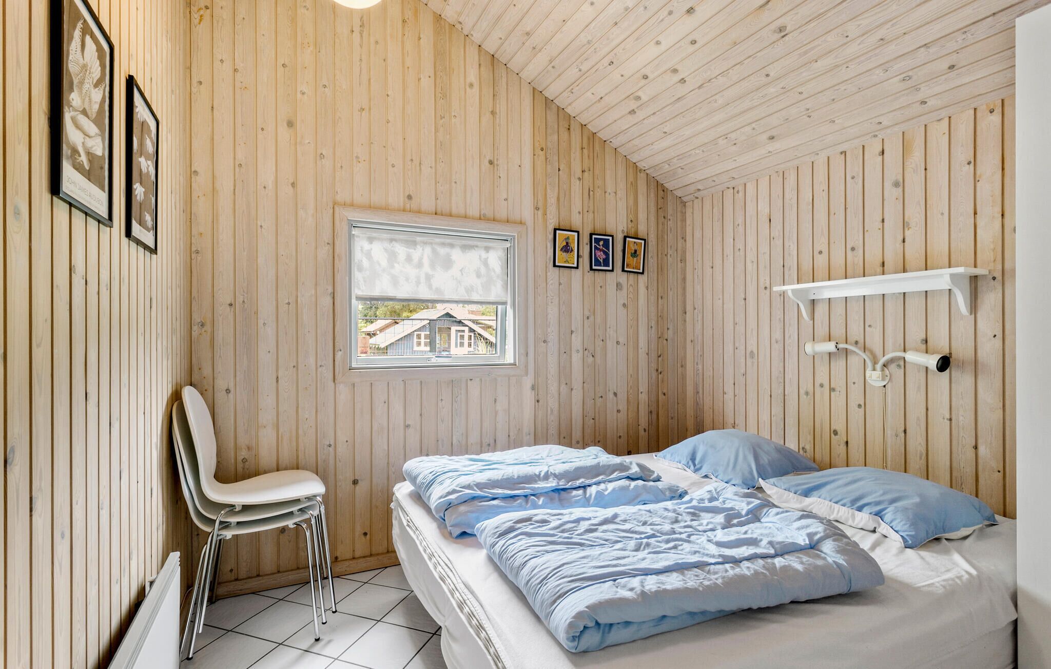 A bedroom with a wooden paneled wall, a bed with blue bedding, a window with a view of a house, and a white shelf with a lamp.