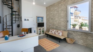 A modern living room with a stone wall, a large window with a view of buildings, a sofa, a TV, and a small staircase. - La Casa in Riva al Mare (Santa Croce Camerina)
