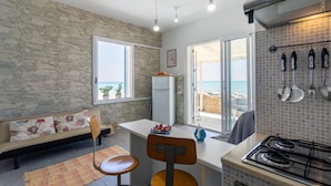 A compact kitchen with a stone wall, a dining area with a bench and chairs, and a view of the sea through large windows. - La Casa in Riva al Mare (Santa Croce Camerina)