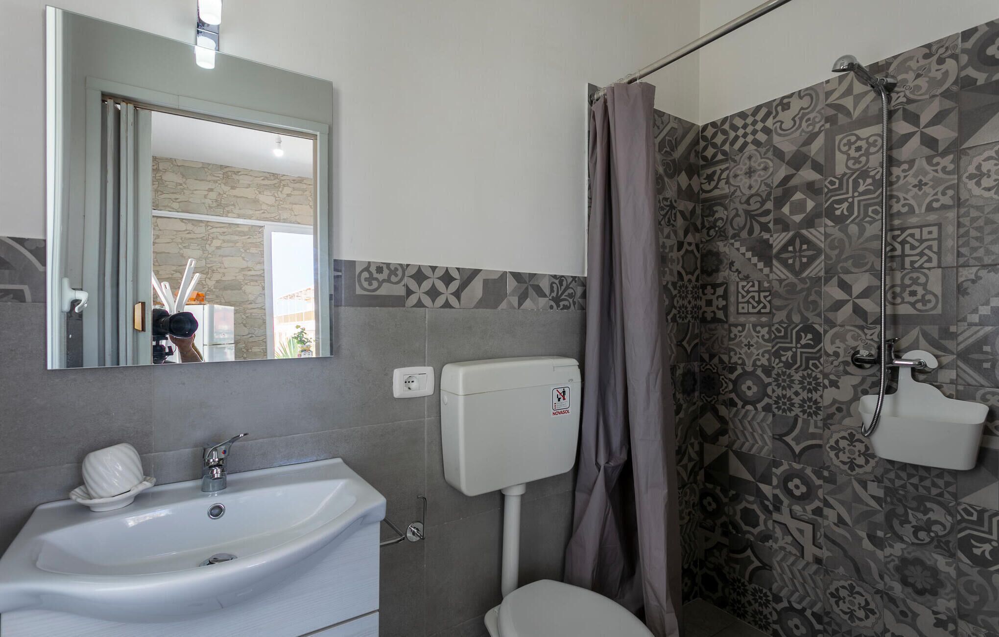 Un baño con un lavamanos blanco, un inodoro, una ducha con azulejos estampados y un espejo que refleja parte del cuarto.
