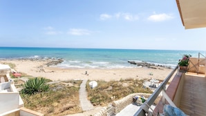 A beach view from a balcony overlooking a sandy beach with a rocky shoreline, a pathway leading to the beach, and a small outdoor seating area. - La Casa in Riva al Mare (Santa Croce Camerina)