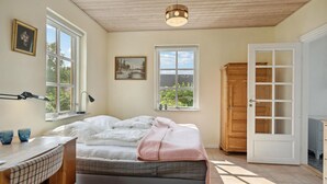 Ein Schlafzimmer mit einem Bett, einem Nachttisch mit Gläsern, einem Schreibtisch, einer Lampe, einem Kleiderschrank und zwei Fenstern.