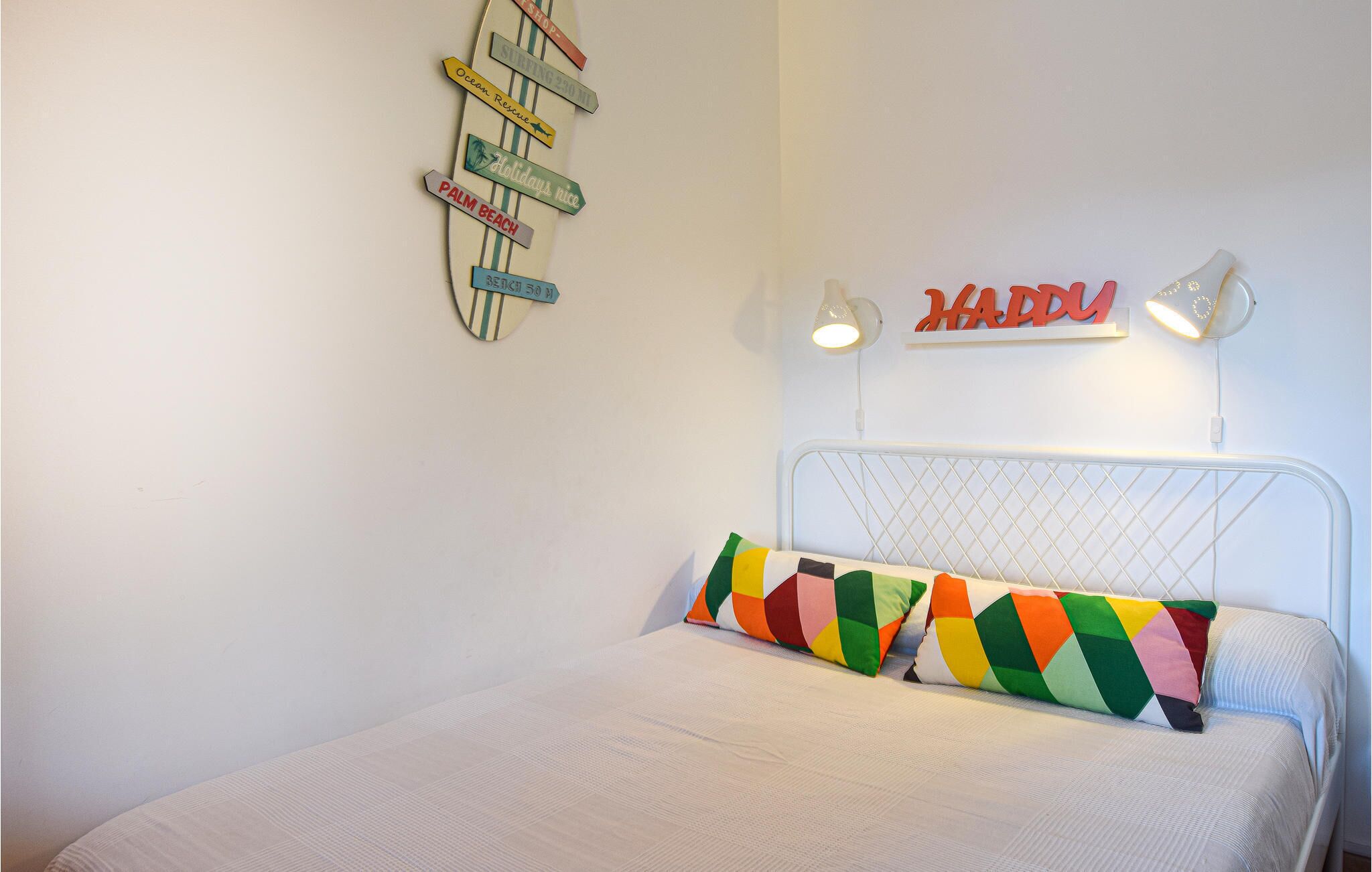 A bedroom with a bed, colorful pillows, a wall-mounted sign with destinations, and two wall-mounted lamps.