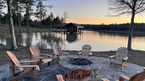 Kivestä rakennettu nuotiopaikka, jonka ympärillä on puisia Adirondack-tyylisiä tuoleja. Taustalla näkyy järvi ja laituri.