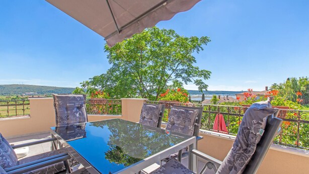 A glass-top table with four chairs, a metal railing, and a view of a hillside and sea.