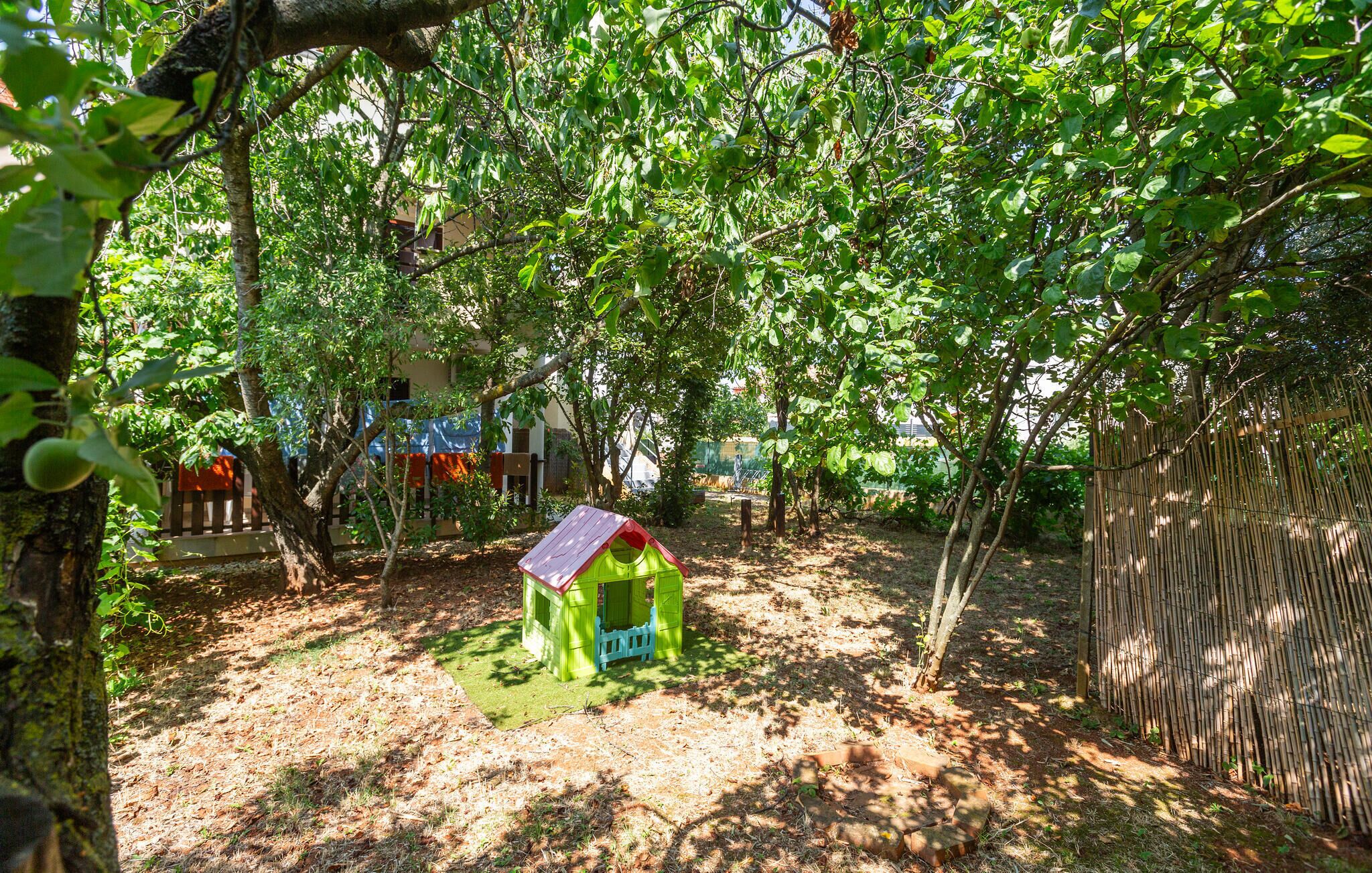 Une petite maison de jeu verte, avec un toit rouge, située dans une zone boisée, entourée d’arbres et d’une clôture en bambou.
