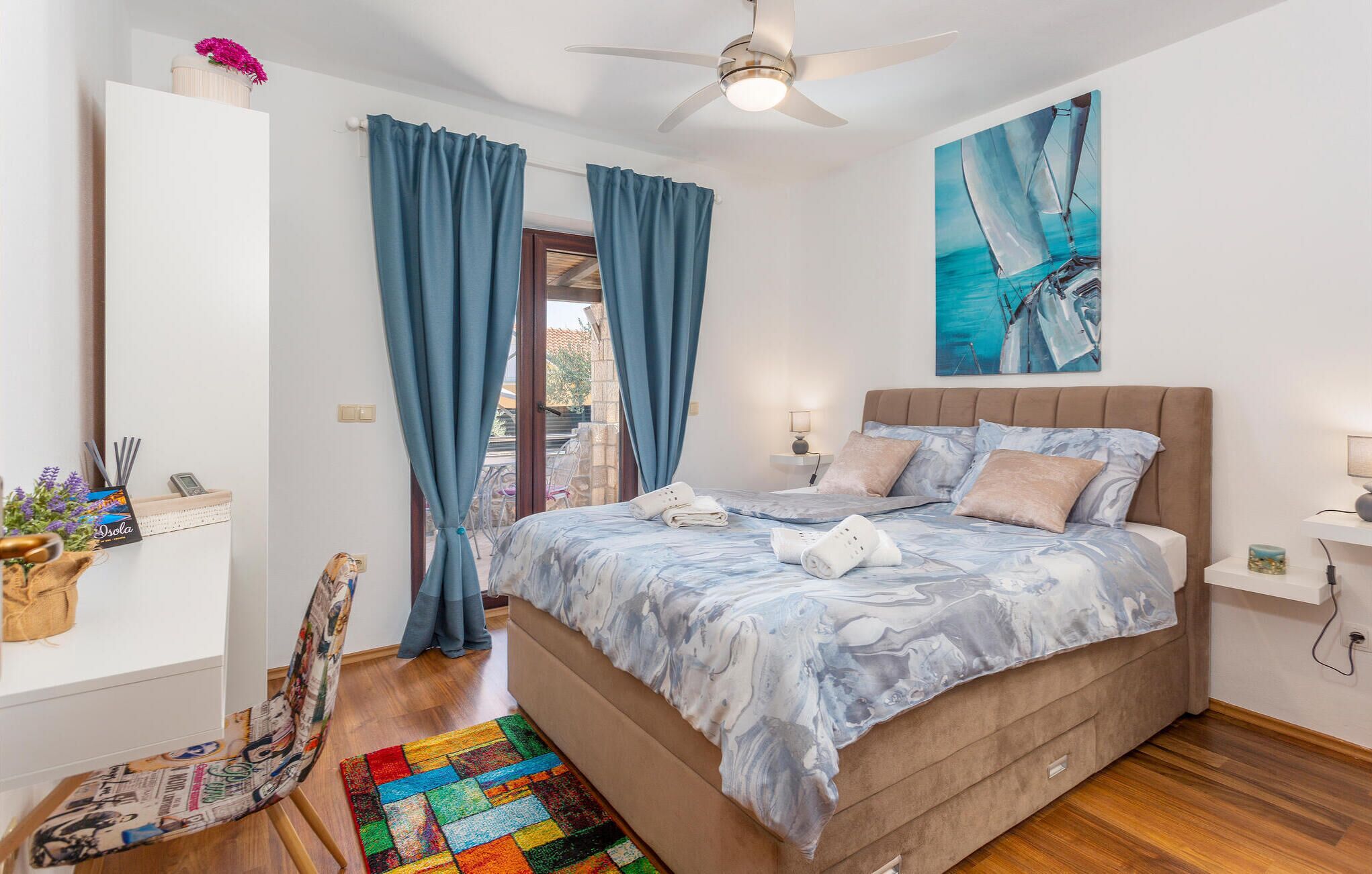 Une chambre à coucher avec un lit, un ventilateur de plafond, un tableau et une fenêtre avec des rideaux.