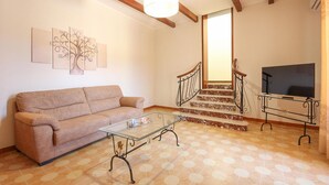 A living room with a sofa, a glass coffee table, a TV on a stand, a staircase with a railing, and a wall with abstract art.