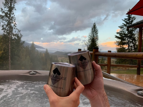 Henkilö pitää kädessään kahta metallista tölkkiä, joissa on merkintä ”Blackridge Resort”. Taustalla näkyy puinen terassi, poreallas ja kaunis näkymä vuorille ja puille pilvisen taivaan alla.