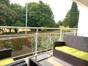 A balcony with wicker furniture, a glass barrier, and a view of a pond and trees.