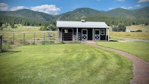 A small, single-story building with a metal roof and white trim, surrounded by a fenced pasture and a paved path leading to it. - Ranch-style home with horse property and fenced yard (Frenchtown)