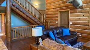 A log cabin interior with a staircase, a black leather sofa, a patterned armchair, and a mounted deer head.