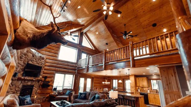 A log cabin interior with a stone fireplace, wooden furniture, and a mounted deer head.