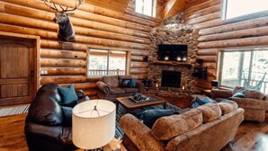 A log cabin interior with a stone fireplace, leather sofas, and a mounted deer head.