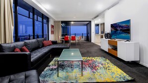 A modern living room with a glass coffee table, black leather sofa, and a colorful rug. There is a TV on a white cabinet and a painting on the wall. - Wave Apartments Broadbeach (Broadbeach)