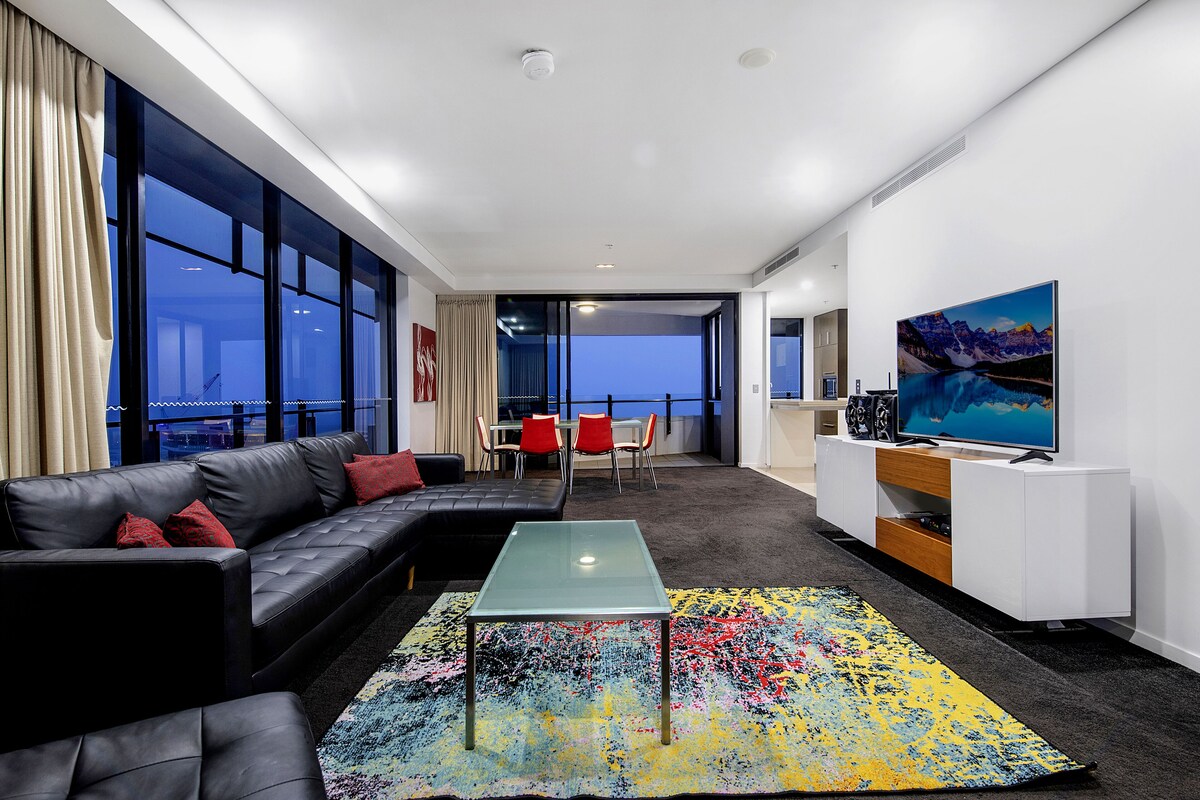 A modern living room with a glass coffee table, black leather sofa, and a colorful rug. There is a TV on a white cabinet and a painting on the wall.