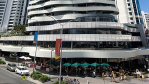 A modern, spiraling building with multiple balconies, surrounded by other high-rise buildings and a street with outdoor seating. - Wave Apartments Broadbeach (Broadbeach)