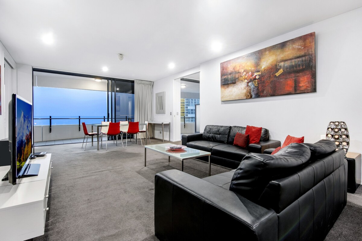 A modern living room with a black leather sofa, a glass coffee table, and a large abstract painting on the wall.