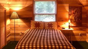Una habitación de cabaña acogedora con techo de madera, colcha a cuadros, dos almohadas con forma de estrella, mesita de noche con lámpara y ventana con persianas.
