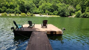Un muelle de madera que se adentra en un lago tranquilo, con dos sillas Adirondack y una caseta pequeña al fondo.