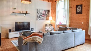 Ein modernes Wohnzimmer mit einem grauen Sofa, einem Fernseher, Holzboden und einem Fenster mit Vorhängen.
