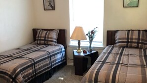 A bedroom with two beds, a nightstand with a lamp, a small table with a vase, and a window with natural light.