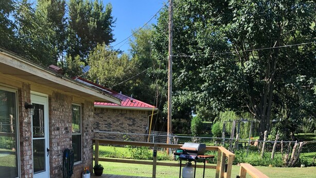 A wooden deck with a metal chair and a table, a brick building with a red roof, and a grassy yard with a wooden fence.