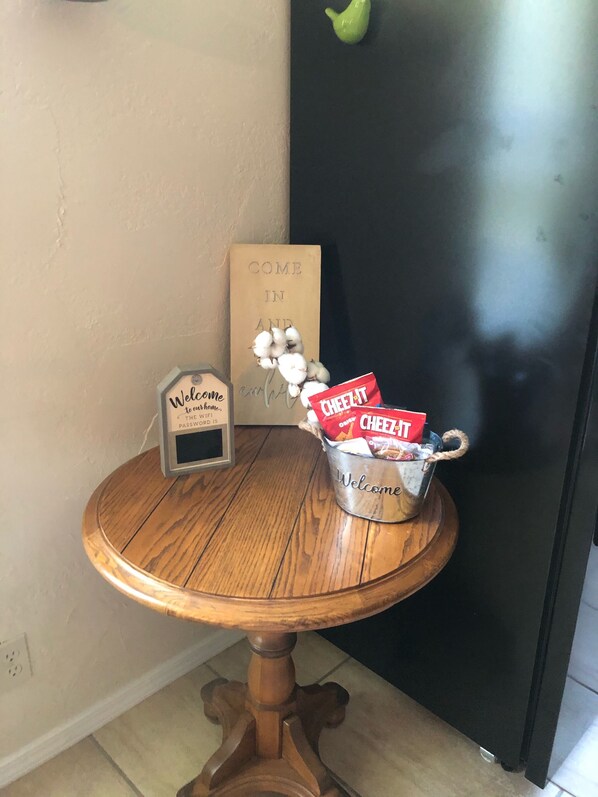 Una pequeña mesa de madera con un cubo metálico que dice 'Bienvenida' y contiene bocadillos, un cartel que dice 'Bienvenidos a nuestro hogar' y un letrero que dice 'Entra y disfruta' en una pared color beige.