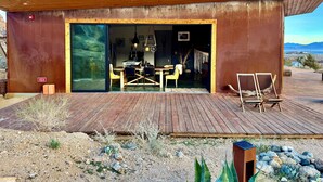 Un bâtiment moderne doté d’une terrasse en bois, d’un coin repas avec des chaises et une table, et offrant une vue sur le paysage.